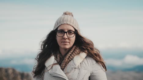 Close-up-shot-of-young-woman-with-glasses-and-long-brown-hair,-lost-in-thought-as-she-gazes-at-the-camera-against-a-stunning-mountain-and-cloudy-sky-backdrop