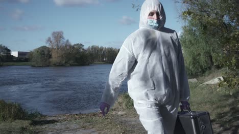 Woman-in-face-mask-and-protective-suit-holding-case-and-walking-out-of-river-or-water,-industrial-background