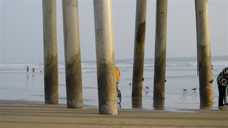 Tall-skinny-caucasian-man-riding-a-bike,-on-the-beach,-under-the-pier