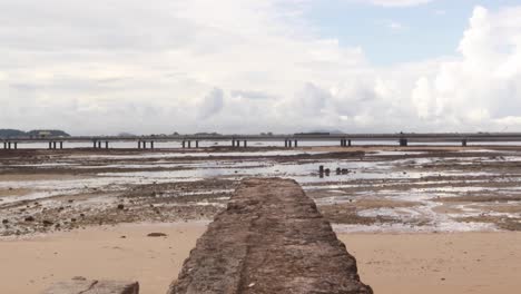An-establishing-tilt-shot-following-a-stone-wall-revealing-the-Cinta-Costera-bridge-which-crosses-the-Panama-Canal,-the-ocean-water-retracting-during-low-tide-leaving-behind-an-empty-bank,-Panama-City
