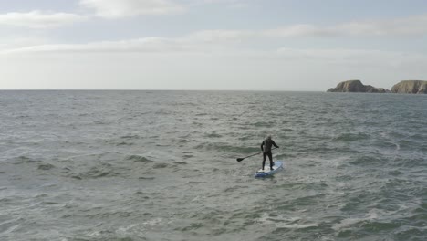Aerial-flyover-of-SUP-paddleboard-male-on-choppy-grey-ocean-near-cliff
