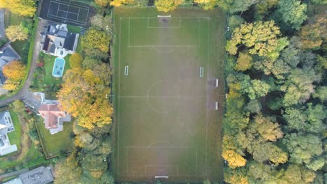 Zoom-out,-top-aerial-view-on-football-field-in-the-middle-of-the-autumn-park-next-to-country-houses-with-swimming-pool-and-tennis-court