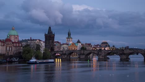Day-to-night-sunset-timelapse-with-a-close-up-view-of-Charles-Bridge-on-Vltava-river-with-boats-on-a-cloudy-evening-in-Prague,-Czech-Republic-as-the-bridge-and-towers-of-the-city-light-up