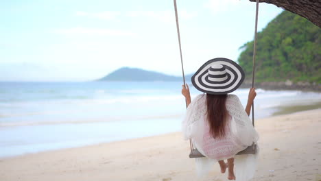 Unrecognizable-Woman-in-Sundress-and-Stripped-Hat-Swinging-on-a-Tree-Swing-with-Ropes-on-Amazing-Sea-Beach-View-Background-in-Thailand-Island---back-view-slow-motion