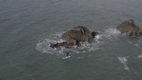 Aerial-of-SUP-athlete-paddling-in-chaotic-water-near-rocky-ocean-islet