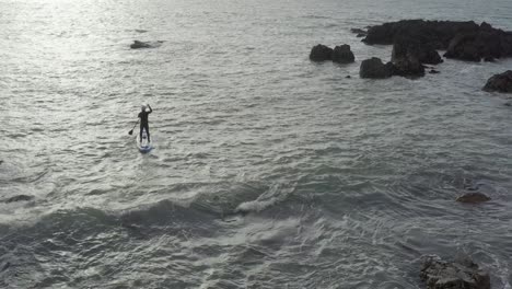 Aerial-follows-male-paddle-boarder-on-choppy-ocean-near-sharp-rocks