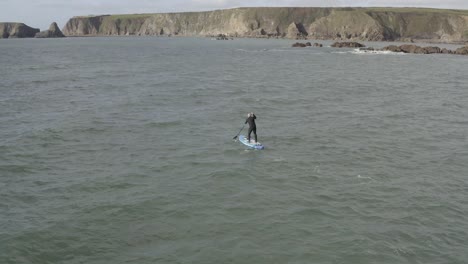 Man-in-wetsuit-turns-paddle-board-toward-rocky-islets-and-sea-cliffs