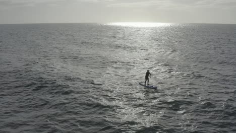 Man-on-stand-up-paddleboard-silhouetted-in-sun-beam-and-ocean-swell