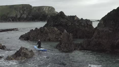 Paddleboarder-in-wetsuit-in-dangerous-ocean-chaos-near-jagged-rocks