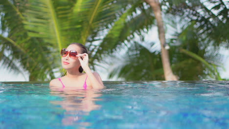 Woman-Touching-Sunglasses-Frame-while-Bathing-in-Swimming-Pool-with-Tropical-Palms-on-Background-in-Thailand-template---slow-motion