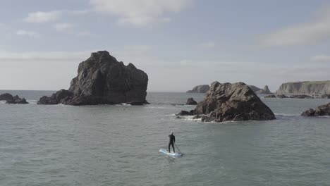 Aerial-follows-SUP-paddleboard-man-on-choppy-ocean-near-jagged-islet