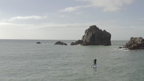 Side-lit-aerial-of-male-SUP-paddle-board-on-sea-near-rugged-rock-islet