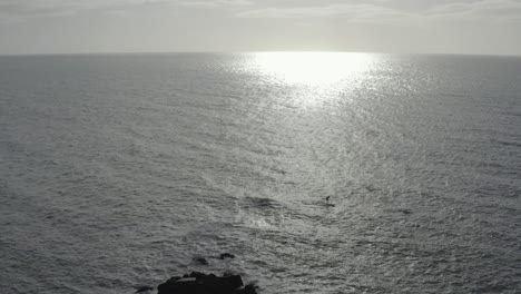 Grey-ocean-aerial-ascends,-retreats-from-SUP-paddleboarder-near-rocks