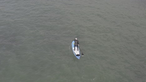 High-angle-aerial-of-wetsuit-man-on-stand-up-paddleboard,-choppy-water