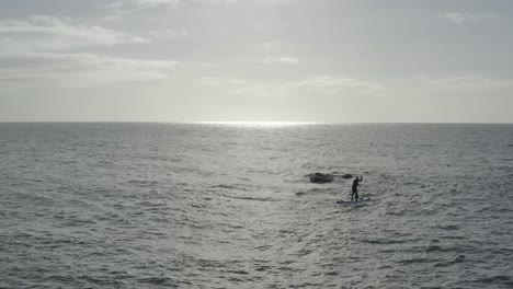 Silhouette-stand-up-paddleboarder-on-flat-grey-ocean-near-rock-hazard