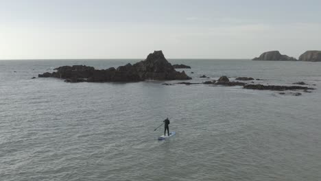 Stand-up-paddleboarder-paddles-toward-rocky-ocean-islets-on-grey-day
