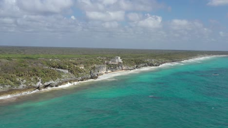 Aerial-forward-over-turquoise-waters-of-Caribbean-sea-and-Mayan-ruins-in-background,-Tulum