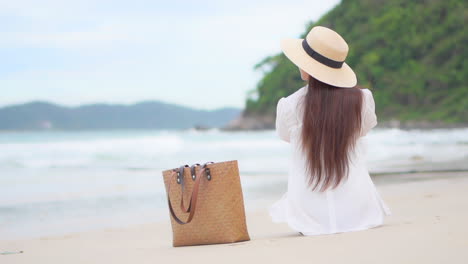 Unrecognizable-woman-sitting-on-a-white-sand-beach-in-front-of-the-sea-wearing-a-white-blouse-shirt-and-straw-hat,-beach-bag,-tropical-Hawaii-island-nature-in-the-background,-slow-motion-back-view