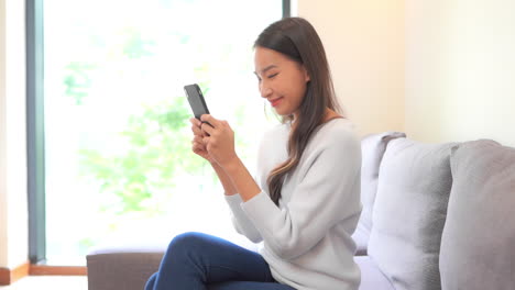 Happy-Thai-asian-woman-using-mobile-phone-holding-it-with-both-hands-while-sitting-on-couch