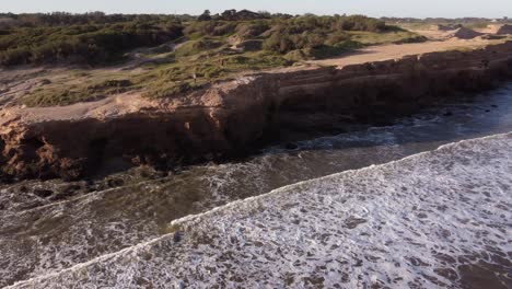 Aerial-view-of-people-walking-on-top-of-big-cliffs-during-sunset-at-Mar-del-Plata-Cliff-in-Argentina---Waves-crashing-against-rocky-wall