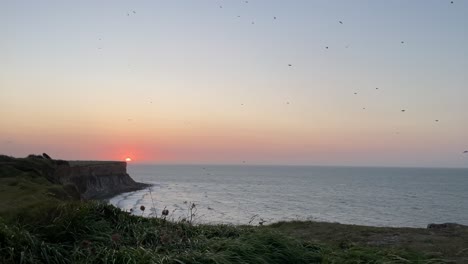 Mystical-sunset-over-cliff-at-D-Day-Landing-Beach,-with-flock-of-birds-in-sky