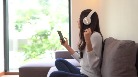 Happy-asian-woman-relaxing-and-using-overhead-headphones-to-listen-to-music-from-smart-phone-sitting-on-sofa-and-snaps-fingers-to-the-beat---side-view
