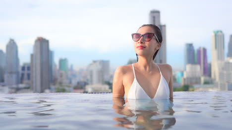 Woman-inside-rooftop-swimming-pool-with-a-view-of-Bangkok-city-skyline-on-sunset,-slow-motion-copy-space