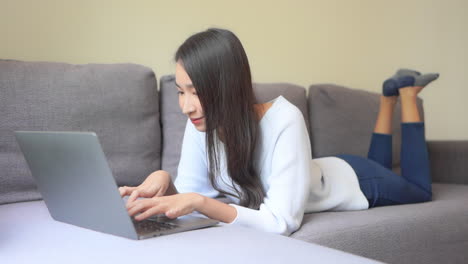 Junge-Asiatische-Frau,-Die-Auf-Ihrem-Laptop-Auf-Dem-Sofa-Tippt