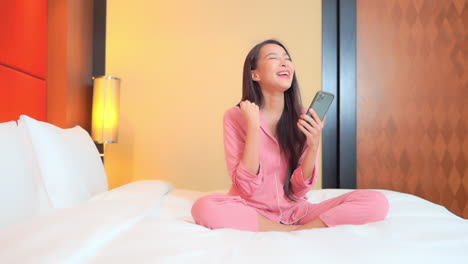A-cute-young-woman-sits-atop-the-comfortable-bed-in-her-hotel-room-as-she-celebrates-a-successful-transaction-on-her-smartphone