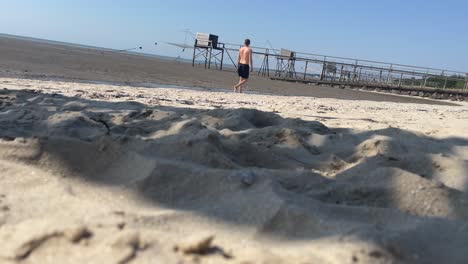 Young-topless-guy-walking-on-beach-in-front-of-famous-fisher-cabines---Baie-de-Bourgneuf-cabanes-de-pêcheurs--at-atlantic-westcoast-in-France,-brittany-in-Pornic
