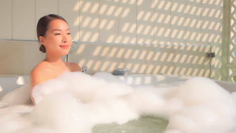 Sexy-Asian-Woman-in-Jacuzzi-Bathtub-Full-of-Foam-Relaxing-With-Sunlight-Coming-From-Window,-Slow-Motion