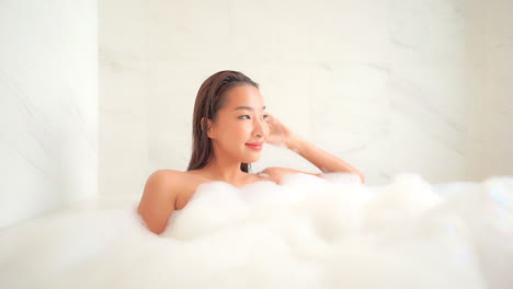 Young-Sexy-Asian-Woman-in-Bathtub-Full-of-Foam-Looking-at-Camera-Relaxing-in-Spa-and-Looking-on-Window,-Slow-Motion,-Full-Frame