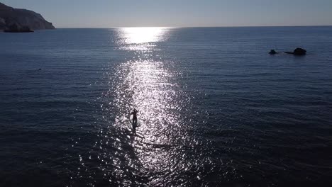 Beautiful-sunrise-aerial-view-of-a-person-paddleboarding-out-to-sea-as-the-sun's-reflection-creates-a-sparkling-path-towards-the-open-water