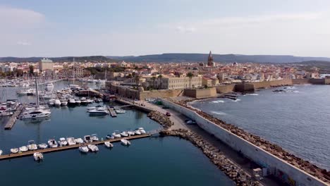 Lateral-drone-clip-over-a-wavebreaker-and-a-marina-with-luxurious-yachts-in-the-old-town-of-Alghero-in-Sardinia,-Italy
