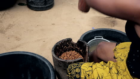 Woman-in-rural-Nigeria,-West-Africa-pounding-tiger-nut-into-flour-in-a-motor-and-pestle---over-shoulder-view