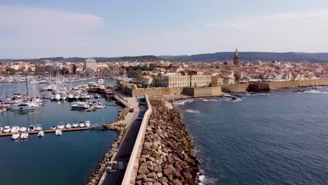 Drone-clip-of-a-luxurious-marina-with-yachts-and-a-wavebreaker-outside-the-castle-walls-of-the-Alghero-town-in-Sardinia,-Italy