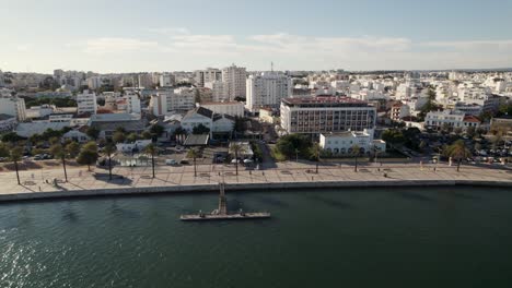 Aerial-orbit-over-Portimão-promenade,-sun-reflecting-on-Arade-river,-CIty-buildings---Algarve