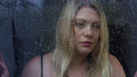 Depressed-blonde-woman-looking-through-a-window-at-the-trees-in-the-rain