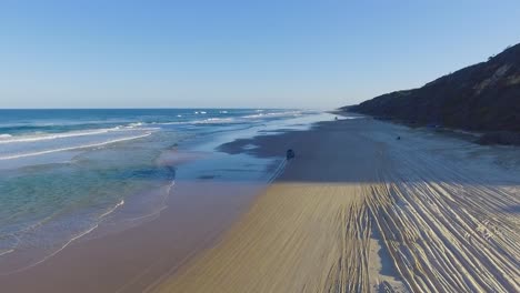 High-quality-and-super-smooth-tracking-shot-at-golden-hour-over-a-broad-Queensland-beach-on-Fraser-Island-with-a-blue-4x4-truck-driving-into-the-distance-past-deep-rutted-tire-tracks
