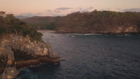 pan-shot-with-drone-from-behind-a-large-rock-in-the-sea-looking-over-a-bay-with-fisherman-boats-at-sunset-on-Nusa-Penida,-Indonesia-in-4k
