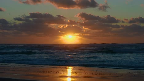 Dramatischer-Blick-über-Einen-Einsamen-Strand-Mit-Dem-Himmel,-Der-Sich-In-Der-Küstenlinie-Spiegelt,-Und-Einem-Atemberaubenden-Goldenen-Sonnenaufgang-Oder-Sonnenuntergang,-Während-Die-Tief-Stehende-Sonne-Von-Der-Rollenden-Und-Schäumenden-Brandung-Funkelt
