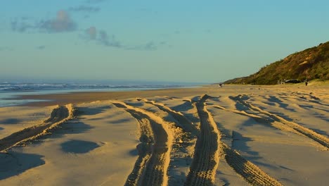 Goldene-Stunde-An-Einem-Strand-In-Queensland-Mit-Tiefen-Schatten-In-Den-Reifenspuren-Der-4x4-trucks-Und-Einer-Sanft-Rollenden-Brandung-Im-Hintergrund
