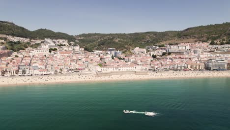 People-enjoying-sand-beach,-Sesimbra-picturesque-townhouses-against-hills-and-blue-sky