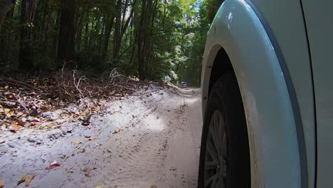 POV-view-past-the-front-wheel-of-a-white-4WD-offroad-truck-from-a-car-mounted-camera,-as-it-bounces-along-a-sandy-inland-trail-through-the-eerie-jungle-rainforest-with-tall-dark-trees-all-around