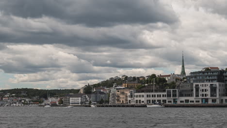Boote-Segeln-Auf-Dem-Ruhigen-Wasser-In-Arendal,-Norwegen-Bei-Bewölktem-Wetter---Stadtansicht-Im-Hintergrund---Zeitraffer