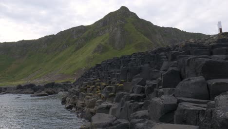 Atemberaubende-Landschaft-Der-Ineinandergreifenden-Sechseckigen-Basaltsäulen-Mit-Den-Grünen-Küstenbergen-Im-Hintergrund-Am-Damm-Des-Riesen-In-Nordirland