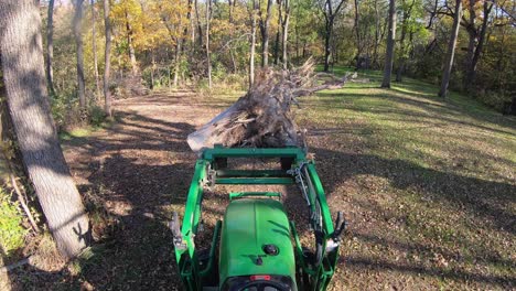 High-angle-point-of-view-on-small-green-tractor-using-lift-forks-to-move-a-tree-stump-along-a-path-in-the-woods-in-autumn