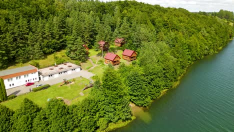 Wooden-Cottage-Houses-with-Red-roofs-for-travelers-accommodation-in-Borucino-forest-near-the-lake,-Poland---pull-back-drone-shot