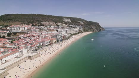 Seagulls-flying-above-the-ocean,-Sesimbra-sand-beach-and-cityscape-against-hills