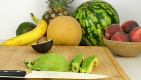 Tracking-shot-of-some-sliced-avocado-on-a-cutting-board,-in-front-of-various-fruit-pieces-and-a-simple-white-background